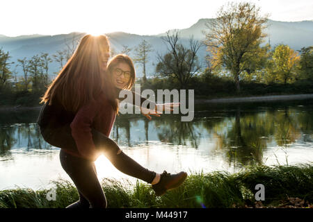 Young woman giving best friend piggy back on riverbank, Calolziocorte, Lombardy, Italy