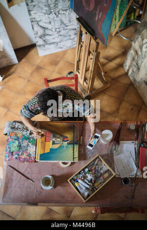 Male artist looking at smartphone while painting canvas in artists studio, overhead view