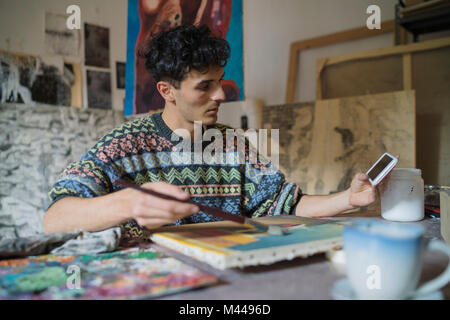 Male artist looking at smartphone while painting canvas in artists studio
