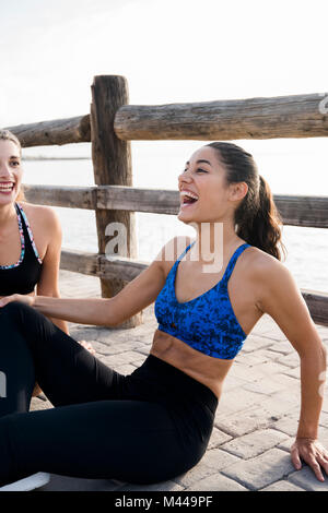 Two young women training on waterfront, sitting laughing Stock Photo