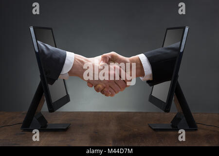 Close-up Of Two Men Shaking Hands Coming Out From Computer Against The Gray Background Stock Photo