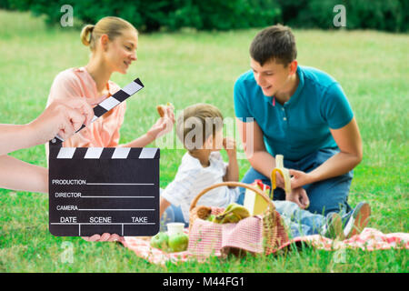 Hands holding clapperboard while young family enjoying picnic on grassy field Stock Photo