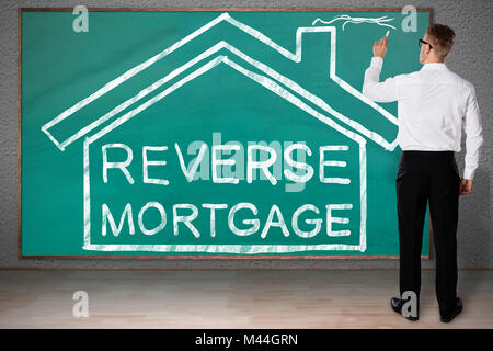 Full length rear view of businessman drawing house with reverse mortgage text on blackboard Stock Photo