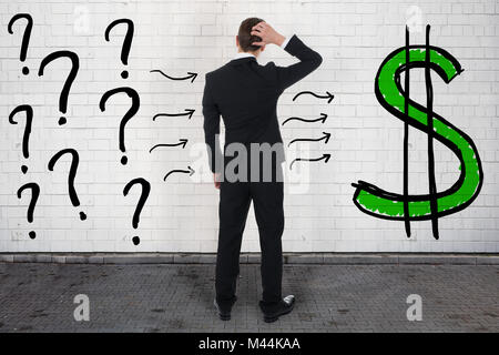 Rear view of confused businessman looking at question marks and dollar sign on wall Stock Photo
