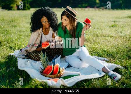 Two multi-ethnic beautiful smiling girl friends are chatting and browsing via the mobile phone while eating the watermelon on the picnic. Stock Photo