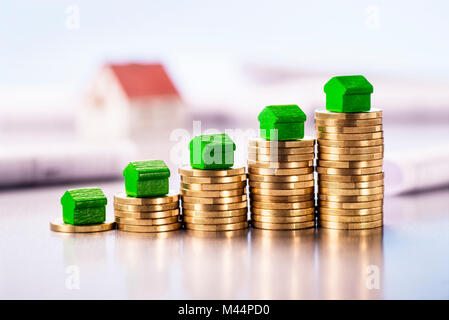 Small green houses standing on stacks of coins with blueprints and architectural model in the background. Stock Photo