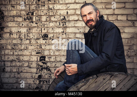 Young bearded man sitting near grungy brick wall, outdoor black and white portrait Stock Photo