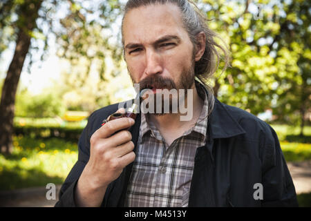 Young serious bearded man smoking pipe in summer park, close-up portrait Stock Photo