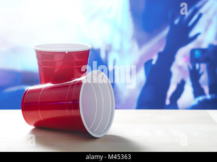 2 plastic red party cups on a table. One on its side. Nightclub or disco full of people dancing on the dance floor in the background. Stock Photo