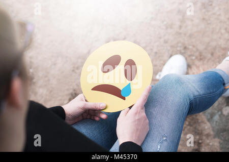 Crying paper emoticon in hand. Depressed woman holding printed sad smiley face and sitting on a rock. Modern communication and smiley icon. Stock Photo