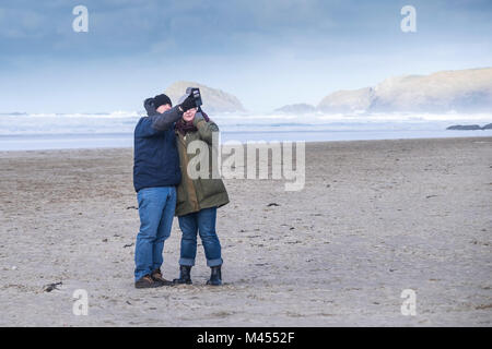 A couple taking a selfie photograph with a smartphone on a windy Perranporth Beach in Cornwall UK. Stock Photo