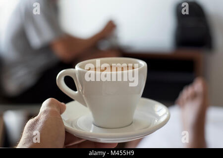 closeup of a young caucasian man with an empty cup of coffee in his hand on bed, and another young man in the background sitting at a table Stock Photo