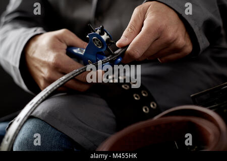 closeup of a young caucasian worker man, wearing a gray working coat, making a hole in a belt with a leather punch Stock Photo