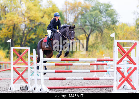 Young rider girl on bay horse jumping over barrier on equestrian sport competition. Horseback girl on show jumping contest Stock Photo