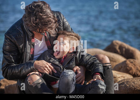 Father and son outdoors, fooling around, sitting on rocks beside sea Stock Photo