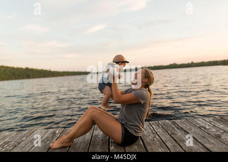 Mother sitting on lake pier holding up baby daughter Stock Photo