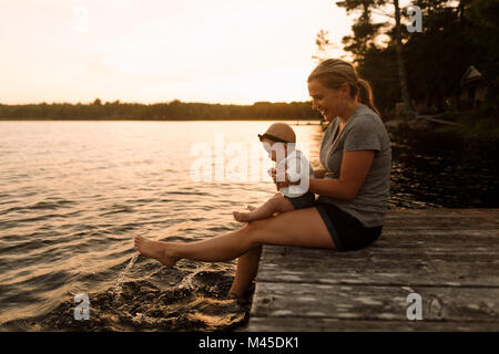 Mother sitting on pier with baby daughter, splashing feet in lake Stock Photo