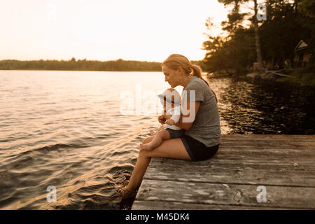 Mother sitting on pier with baby daughter looking down at  lake Stock Photo