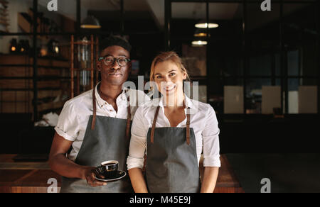Happy coffee shop owner couple standing inside their shop. Man and woman baristas standing inside their café wearing apron with the man holding coffee Stock Photo