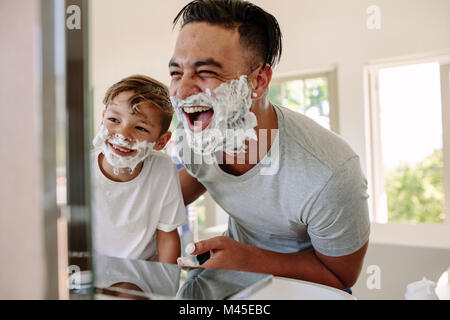 Happy father and son having fun while shaving in bathroom. Young man and little boy with shaving foam on their faces looking into the bathroom mirror  Stock Photo