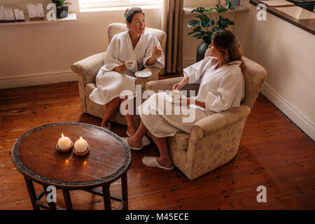 Two women talking while waiting for spa. Female friends in bathrobe sitting on chair having herbal tea and chatting. Stock Photo