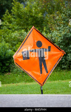 Closeup of an orange, diamond-shaped, men working warning sign standing on the side of a road. The silhouette of a human holding a flag is printed on  Stock Photo