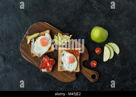 top view of toasts with fried eggs for breakfast on wooden cutting board on dark surface Stock Photo