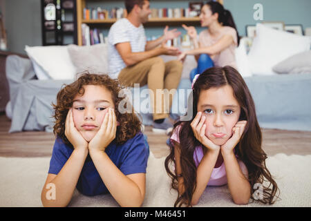 Sad siblings lying on carpet while parents sitting in background Stock Photo