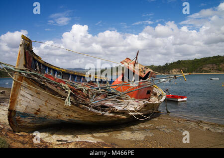 Old fishing boat turned on a side, wrecked and rotting on a rocky shore by the Mira River bank. Bright blue clouded sky. Vila Nova de Milfontes, Portu