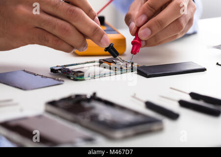 Close-up Of A Technician's Hand Examining Mobile Phone With Digital Multimeter On Desk Stock Photo