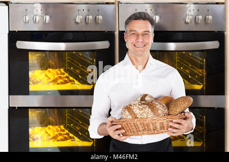 Smiling Baker Holding A Wicker Basket Full Of Bread In Front Of Oven Stock Photo