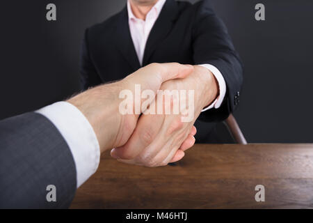 Close-up Of A Two Businessmen Shaking Hands On Wooden Desk Against Gray Background Stock Photo