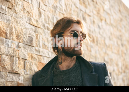 Close up portrait of a stylish bearded man wearing sunglasses and coat leaning on a wall outdoors Stock Photo