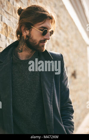 Portrait of a confident bearded man wearing sunglasses and coat leaning on a wall outdoors Stock Photo