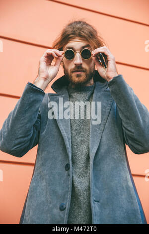 Portrait of a stylish bearded man wearing sunglasses and coat standing against a wall on a city street Stock Photo