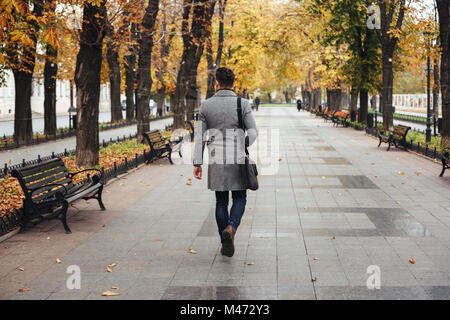 Portrait from back of young stylish guy in coat with bag strolling in city park looking on colorful trees Stock Photo