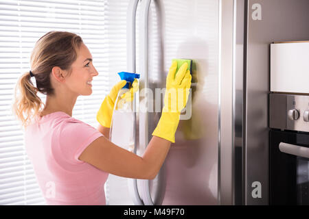 Happy Young Woman Cleaning Stainless Refrigerator With Sponge And Bottle Spray In Kitchen Stock Photo