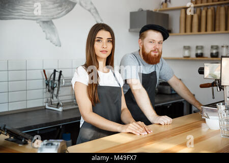 Coffee Business Concept - Positive young bearded man and beautiful attractive lady barista couple in apron looking at camera while standing at bar Counter Stock Photo