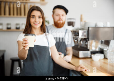 Coffee Business Concept - Positive young bearded man and beautiful attractive lady barista couple giving take away cup of coffee to custome at the modern coffee shop Stock Photo