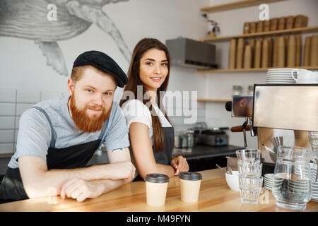 Coffee Business Concept - Positive young bearded man and beautiful attractive lady barista couple in apron looking at camera while standing at bar Counter Stock Photo