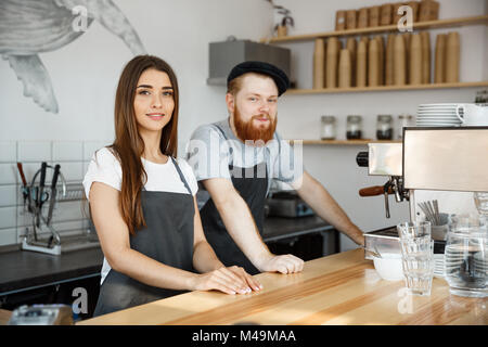 Coffee Business Concept - Positive young bearded man and beautiful attractive lady barista couple in apron looking at camera while standing at bar Counter. Stock Photo