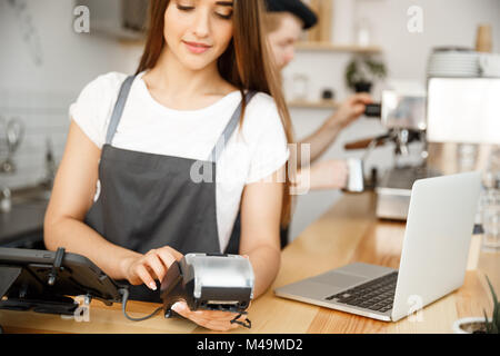 Coffee Business Concept - Beautiful female barista giving payment service for customer with credit card and smiling while working at the bar counter in modern coffee shop. Stock Photo