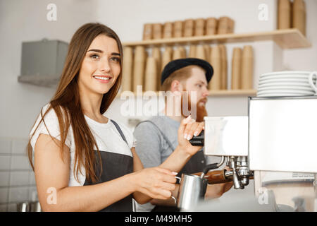 Coffee Business Concept - portrait of lady barista in apron preparing and steaming milk for coffee order with her partner while standing at cafe. Stock Photo