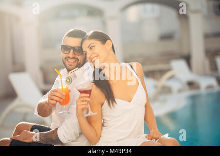 Youbg cheerful loving couple having cocktails by the pool on summer vacation.Romantic evening in luxury resort.Celebrating love ,engagement,anniversar Stock Photo