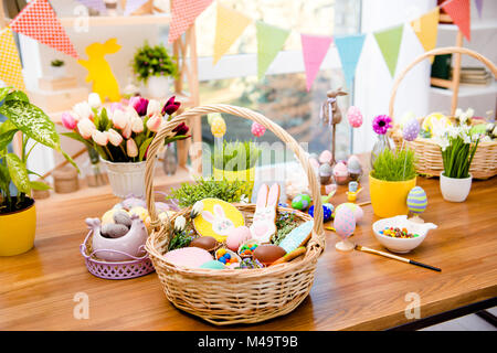 Wooden basket with easter composition, sweets, choco, gingerbread standing on table decorated with easter colored eggs and flowers Stock Photo