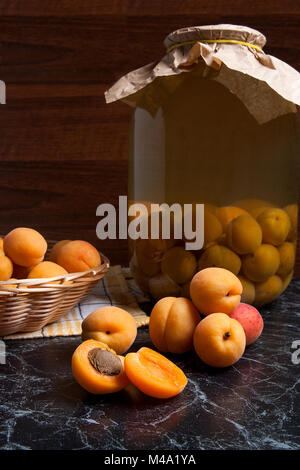 Ripe organic apricots harvested at summer time and big glass jar with canned sweet apricot compote on dark marble. Composition in rustic style - organ Stock Photo