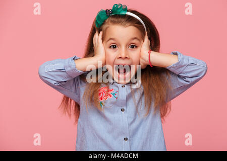 Surprised little girl covering her ears with both hands not listening or overhearing, screaming with open mouth over pink background Stock Photo