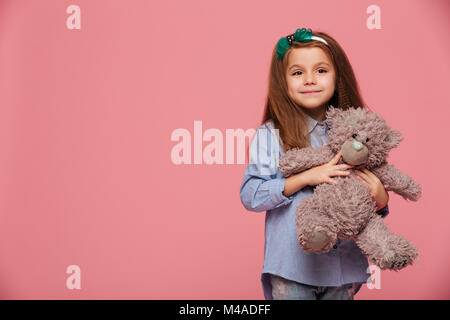 Image of sweet schoolgirl 5-6 years with long auburn hair smiling, holding her lovely teddy bear isolated over pink background Stock Photo