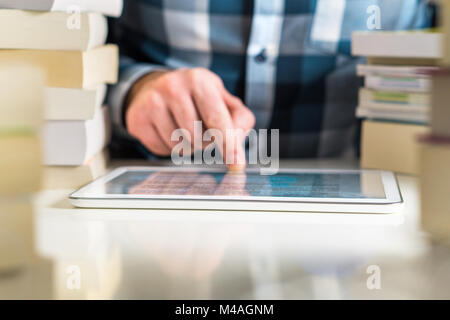 Man reading electronic book with reader. Touching screen with finger. Book on tablet or smart mobile device surrounded by stacks of books. Stock Photo