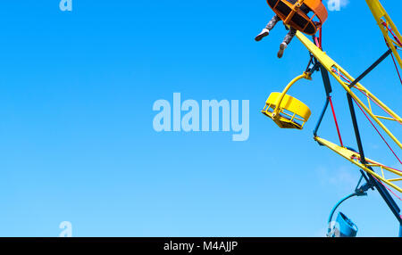 Amusement park background template with negative copy space. Colorful theme park ride against clear blue sky in summer. One person with legs hanging. Stock Photo